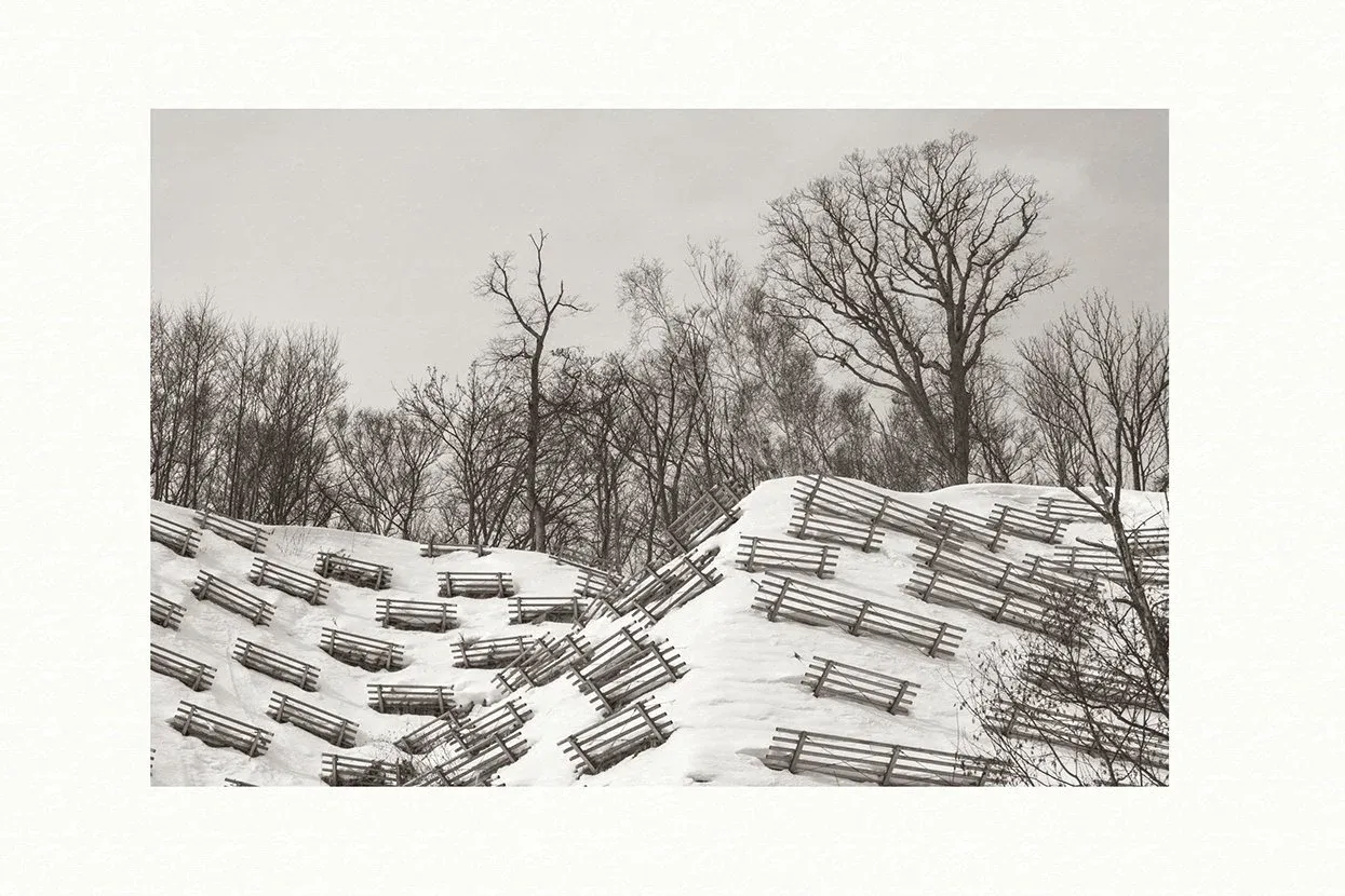 Tirage noir et blanc aux tons chauds sur Awagami Bizan : rangées de barrières à neige en bois sur une colline enneigée, arbres nus sous un ciel gris
